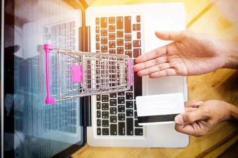 Person holding a credit card near a laptop with a miniature shopping cart, symbolizing online shopping or eCommerce transactions.