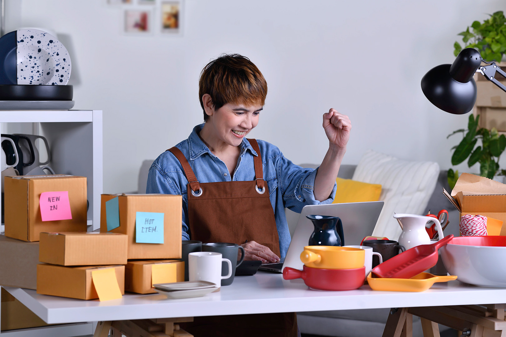 rjvgv8hxwbr4gbvyj6ws (1) Happy female entrepreneur celebrating success while working on a laptop in a home office, surrounded by ceramic products and shipping boxes labeled.<br />